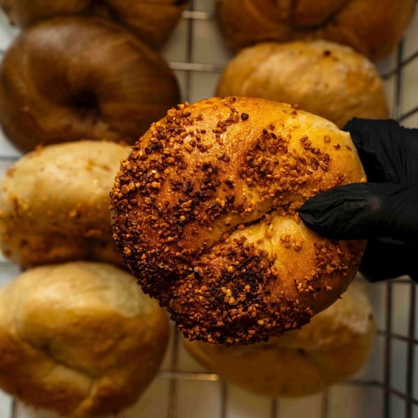 A gloved hand holds a freshly baked Nana's Bagel with a crunchy topping, surrounded by other bagels.