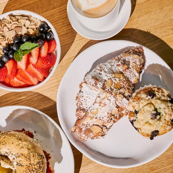 Overhead view of Nana's Bagel breakfast spread: bagel sandwich, fruit bowl, pastry, muffin, and latte.