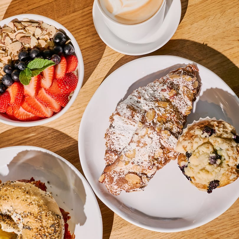 Overhead view of Nana's Bagel breakfast spread: bagel sandwich, fruit bowl, pastry, muffin, and latte.