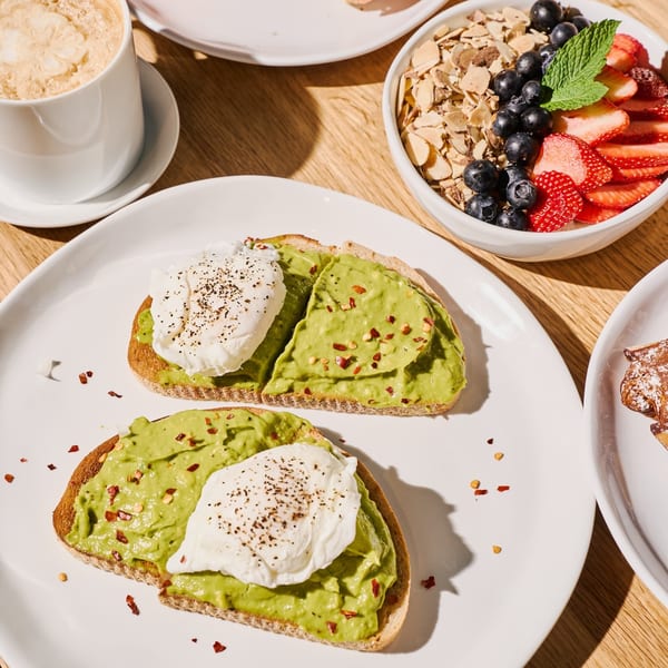 Avocado toast with poached eggs and chili flakes, alongside a fruit and granola bowl and coffee.