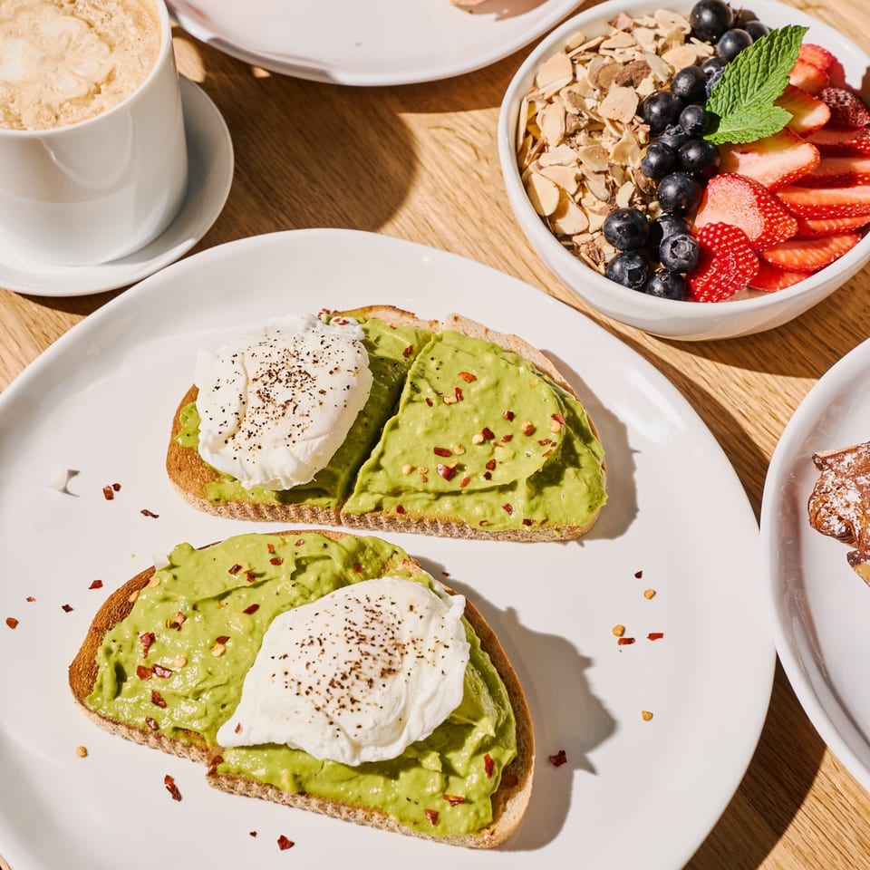 Avocado toast with poached eggs and chili flakes, alongside a fruit and granola bowl and coffee.